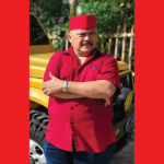 Karl Sibarani mengenakan kemeja merah dan peci merah, berdiri dengan tangan terlipat di depan mobil berwarna kuning, saat menyampaikan pandangannya tentang fenomena sosial terkait polemik beasiswa negara dan krisis identitas kebangsaan.