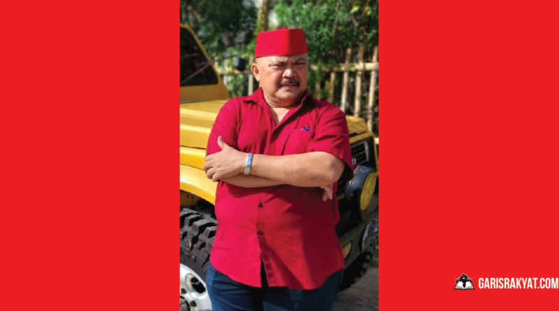 Karl Sibarani mengenakan kemeja merah dan peci merah, berdiri dengan tangan terlipat di depan mobil berwarna kuning, saat menyampaikan pandangannya tentang fenomena sosial terkait polemik beasiswa negara dan krisis identitas kebangsaan.